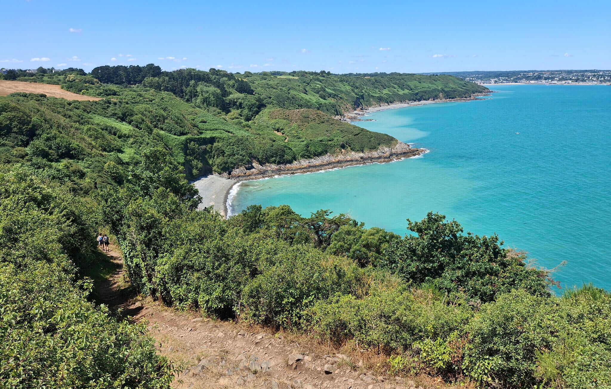 A coastal landscape with a beach, dense greenery, and a clear blue sea.