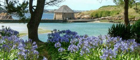 A stone building with a conical roof by a calm body of water, surrounded by lush greenery and purple flowers.
