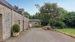 Un bâtiment en pierre avec une allée de gravier menant à une table et à des bancs en bois, le tout entouré de verdure.