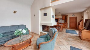Un salon comprenant un canapé, un fauteuil, une table basse en verre, une cheminée, un coin cuisine et un téléviseur.