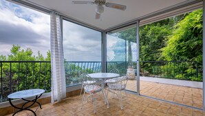 A balcony with a table and chairs, large windows, and a ceiling fan. - Charmante villa vue mer (Grasse Magagnosc)
