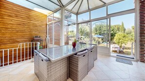 Eine überdachte Terrasse mit Glastisch und Korbmöbeln, mit Blick auf einen Garten.