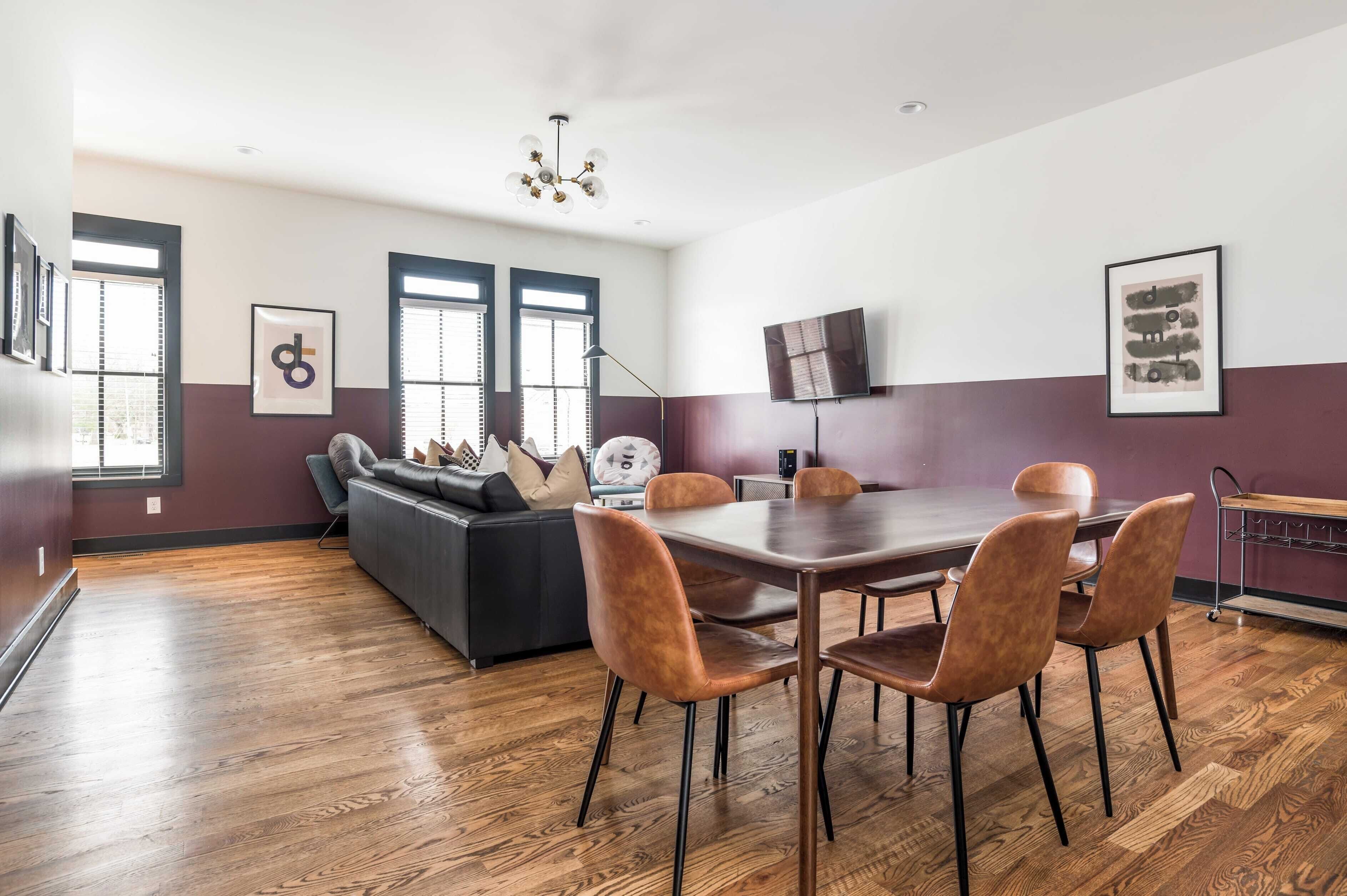 A modern dining area with a wooden table, four chairs, a sofa, and a TV mounted on the wall.