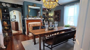 Un coin repas avec une table en bois, des chaises et un lustre. On y trouve une cheminée avec un manteau et divers objets décoratifs. La pièce est dotée d’un parquet en bois et d’un grand miroir au mur.