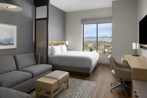 A modern hotel room with a bed, sofa, desk, and a large window offering a city view.