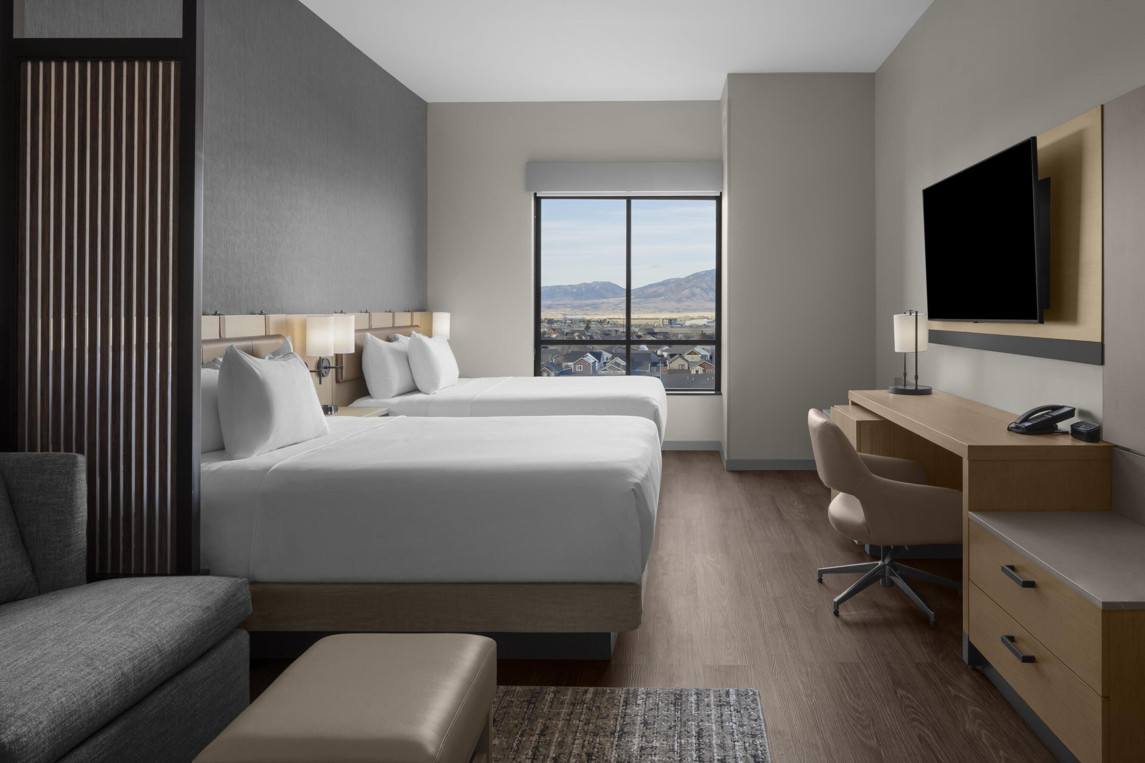 A hotel room with two beds, a desk, a chair, a television, and a large window with a city view.
