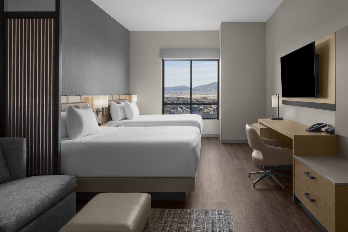 Habitación de hotel moderna con dos camas, un escritorio, una silla, un televisor y un amplio ventanal con vistas a la ciudad.
