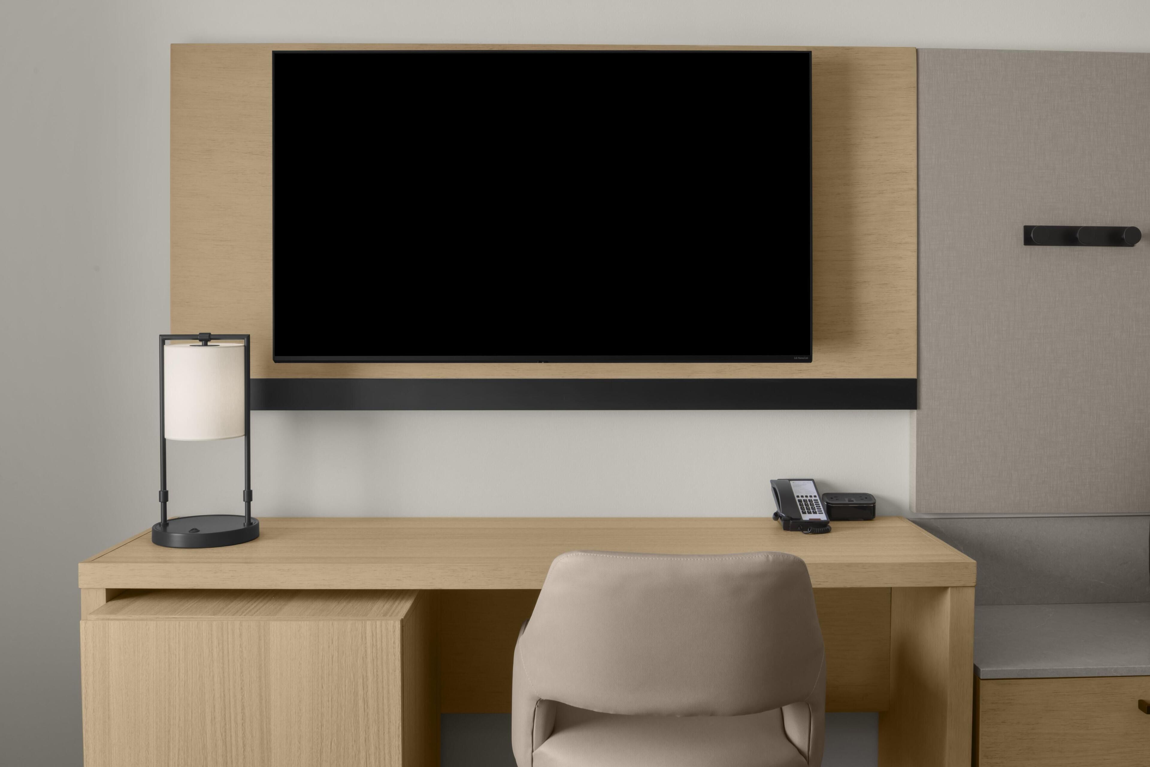 Un espace de bureau moderne comprenant un bureau, une chaise, un téléviseur fixé au mur, une lampe, un téléphone et un petit meuble de rangement.
