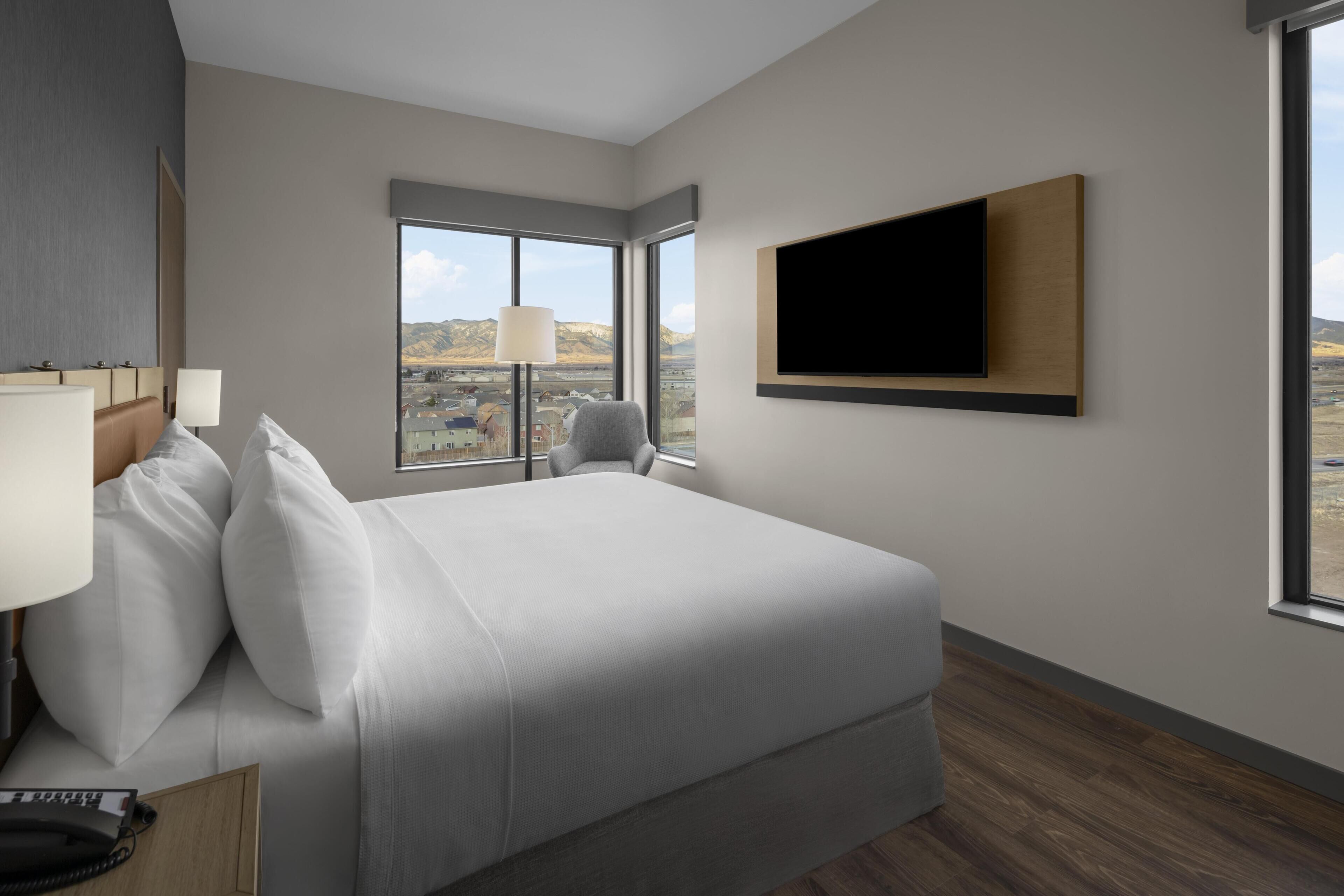 A hotel room with a large bed, a TV mounted on the wall, a desk with a lamp, and a view of the mountains through the window.
