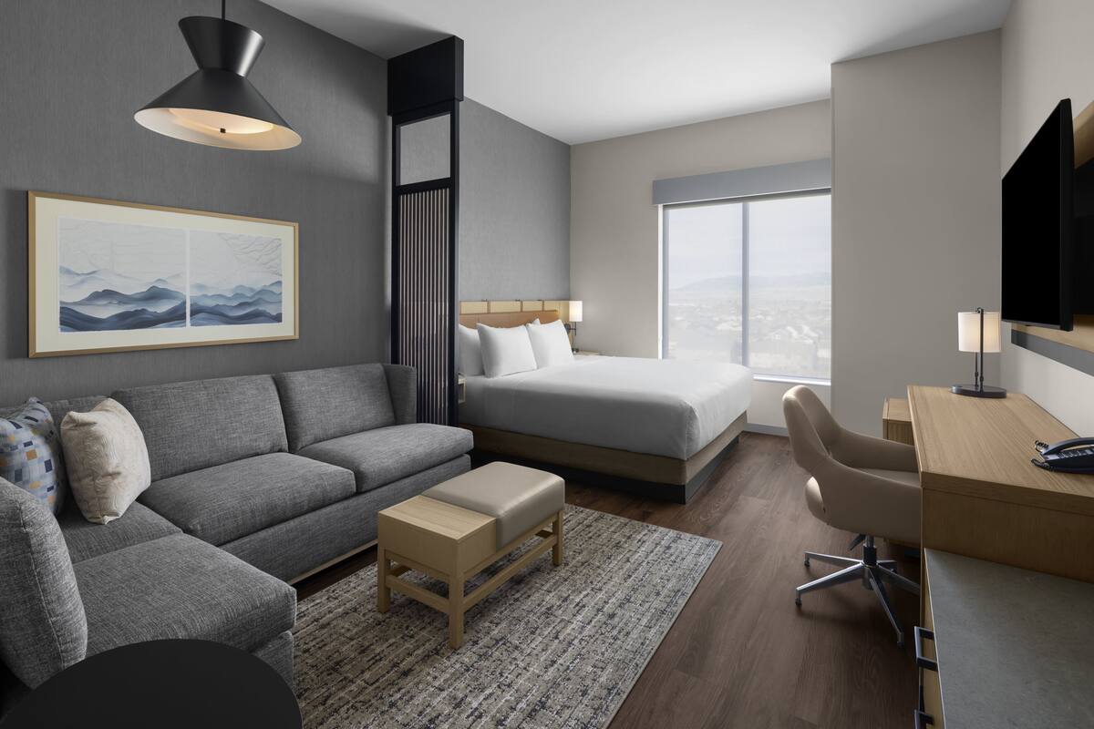 Una habitación de hotel moderna con un sofá gris, una cama, un escritorio y un ventanal grande.