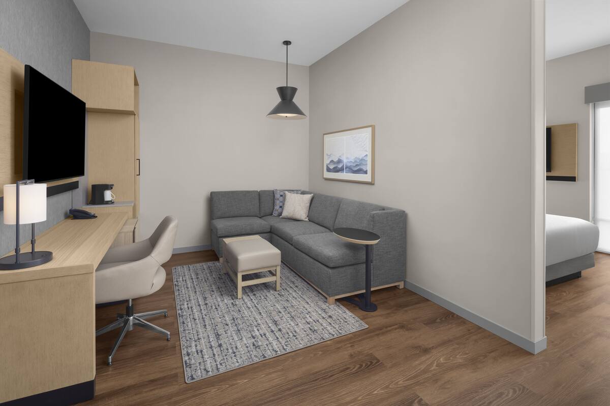 Una habitación de hotel moderna con un sofá gris, una mesita redonda pequeña, un escritorio con una silla, un televisor montado en la pared y un cuadro enmarcado.