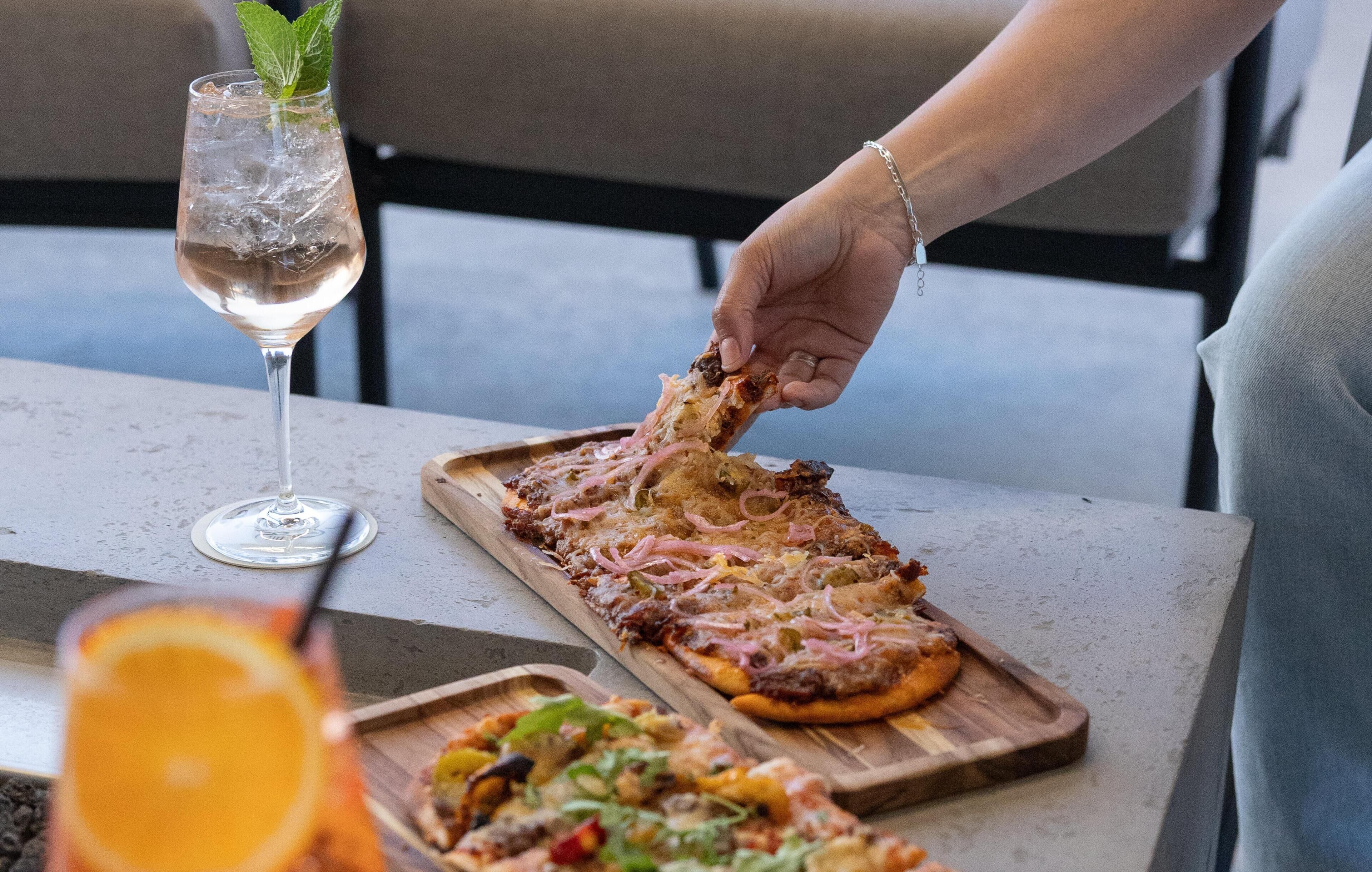 A person holding a slice of pizza with toppings, a glass of a clear beverage with ice, and a wooden serving tray on a table.
