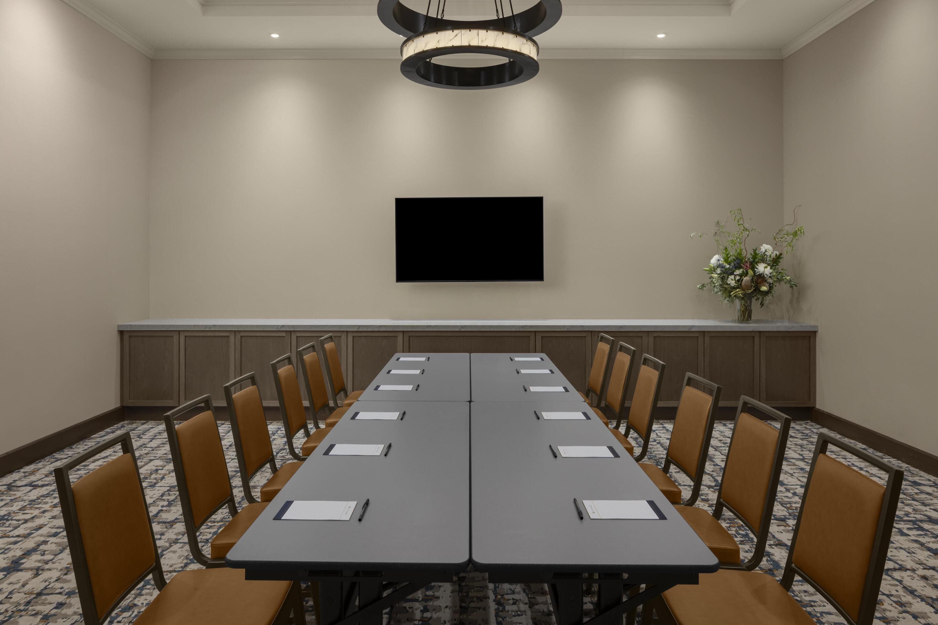 A conference room with a long table, chairs, a flat-screen TV, and a vase with flowers.
