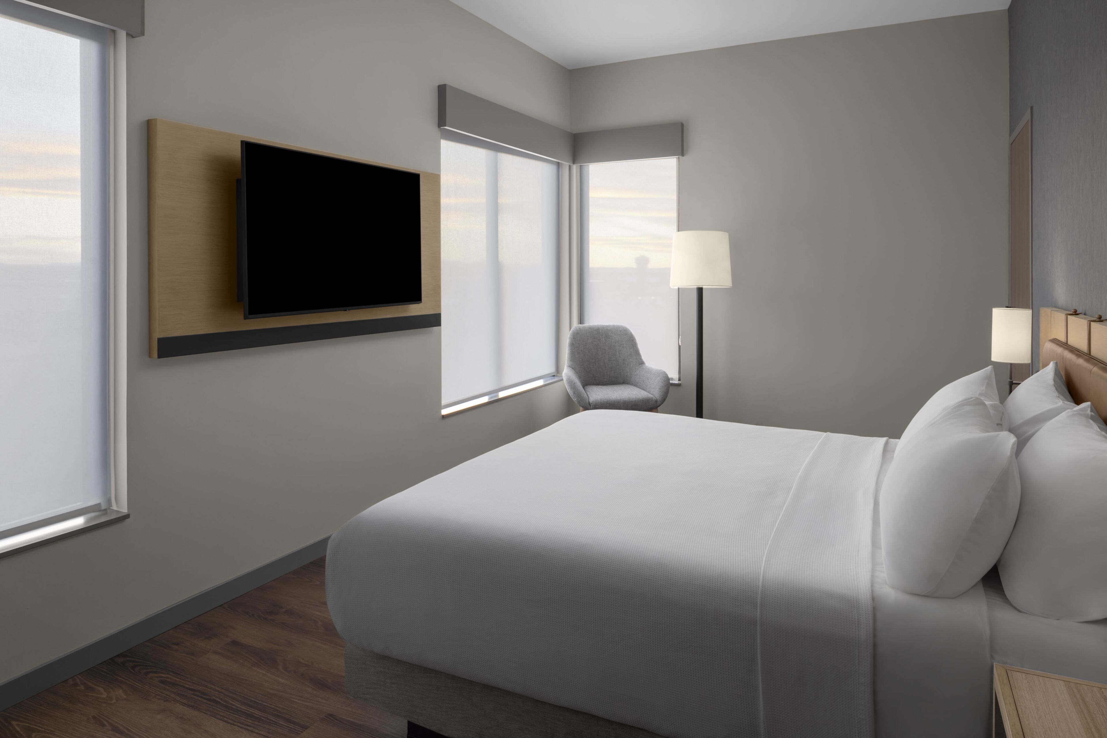 Una habitación de hotel moderna con una cama grande, un televisor de pantalla plana montado en la pared, una silla, una mesita de noche con lámparas y ventanas amplias.