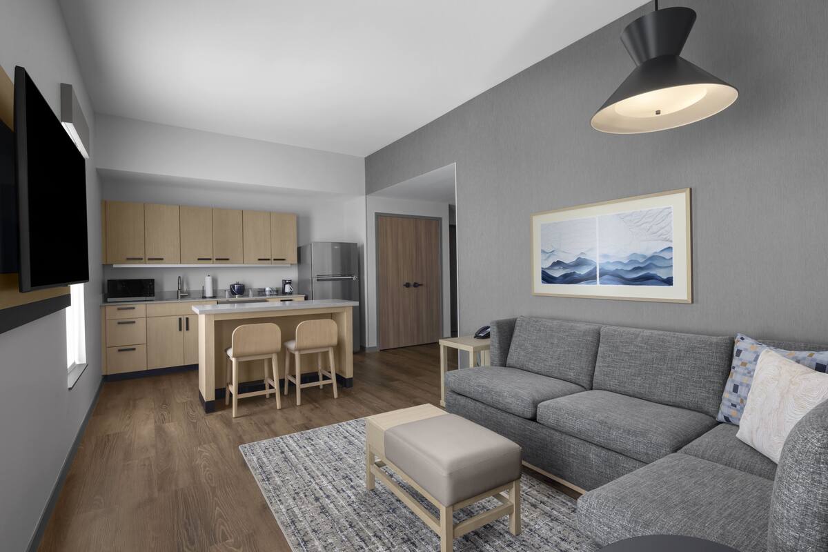 Una sala de estar moderna con un sofá gris, una mesita, un televisor montado en la pared y una cocina al fondo.