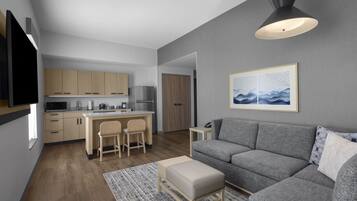 Un salon moderne avec un canapé gris, une petite table, un téléviseur fixé au mur et un coin cuisine à l’arrière-plan.