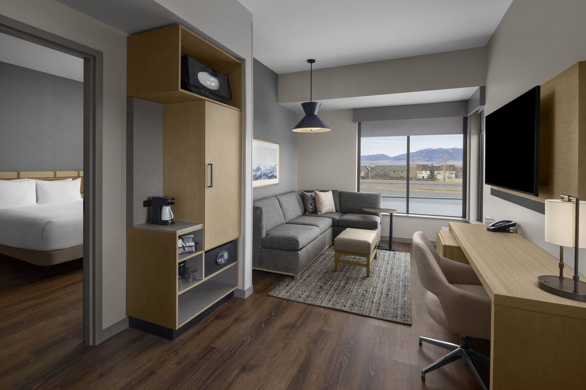 Habitación de hotel moderna con cama, sofá, escritorio, televisor y ventana grande con vistas a la montaña.