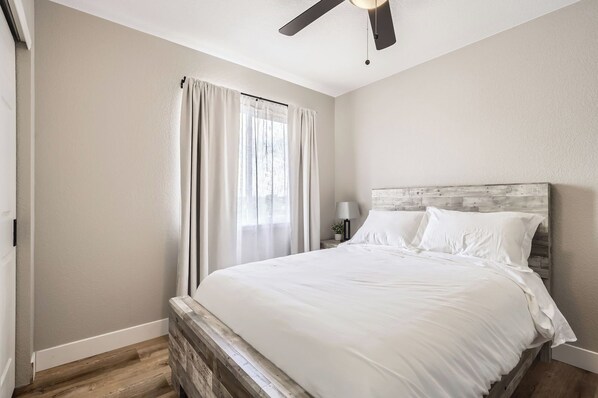 A bedroom with a bed, a ceiling fan, a nightstand with a lamp, and a window with curtains.