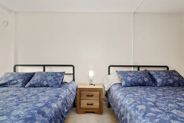 A bedroom with two beds, each with a floral-patterned duvet, a bedside table with a lamp, and a wooden drawer unit.