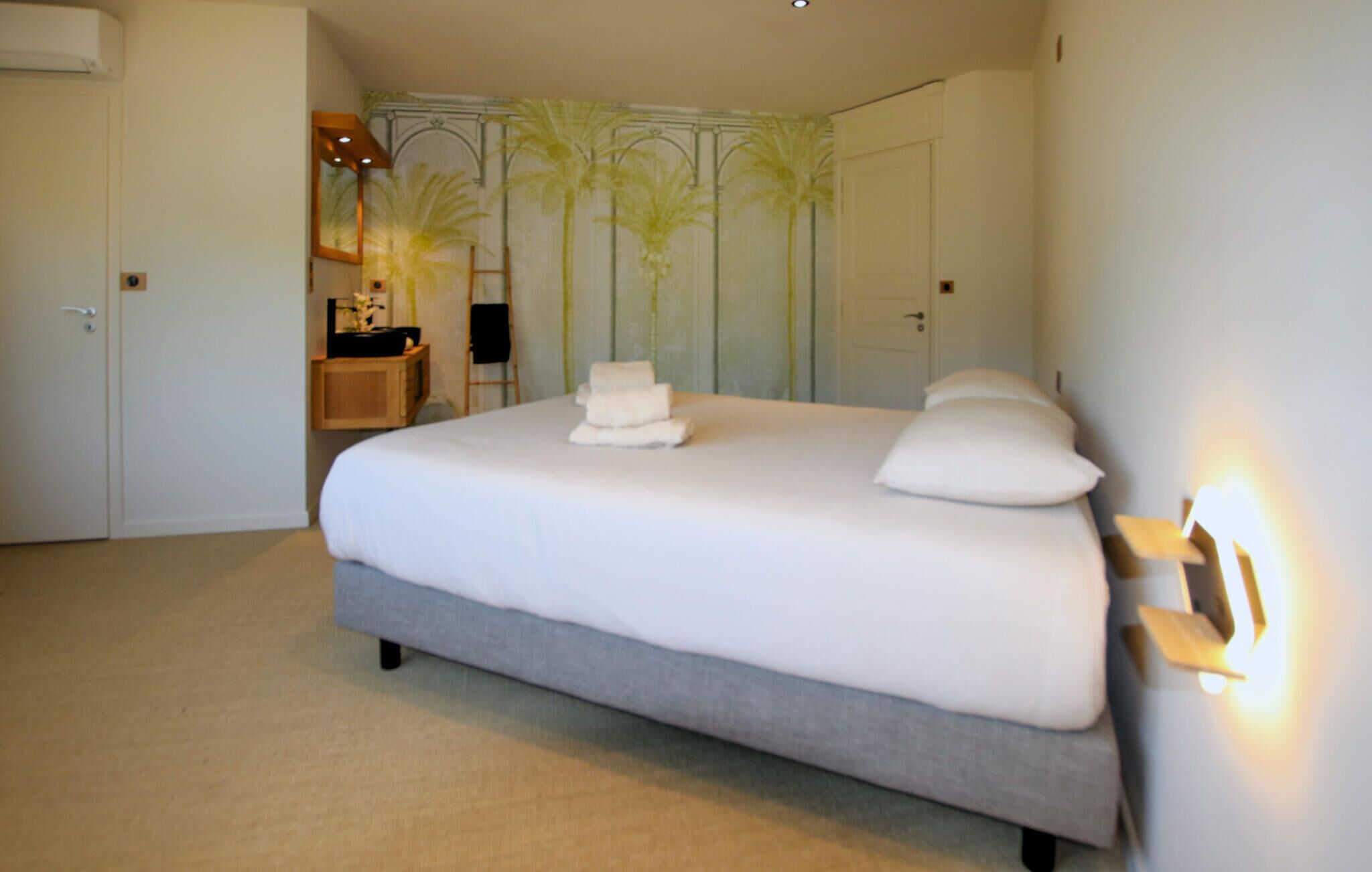 A neatly made bed with white linens and pillows, a bedside table with a lamp, and a wall with palm tree wallpaper.
