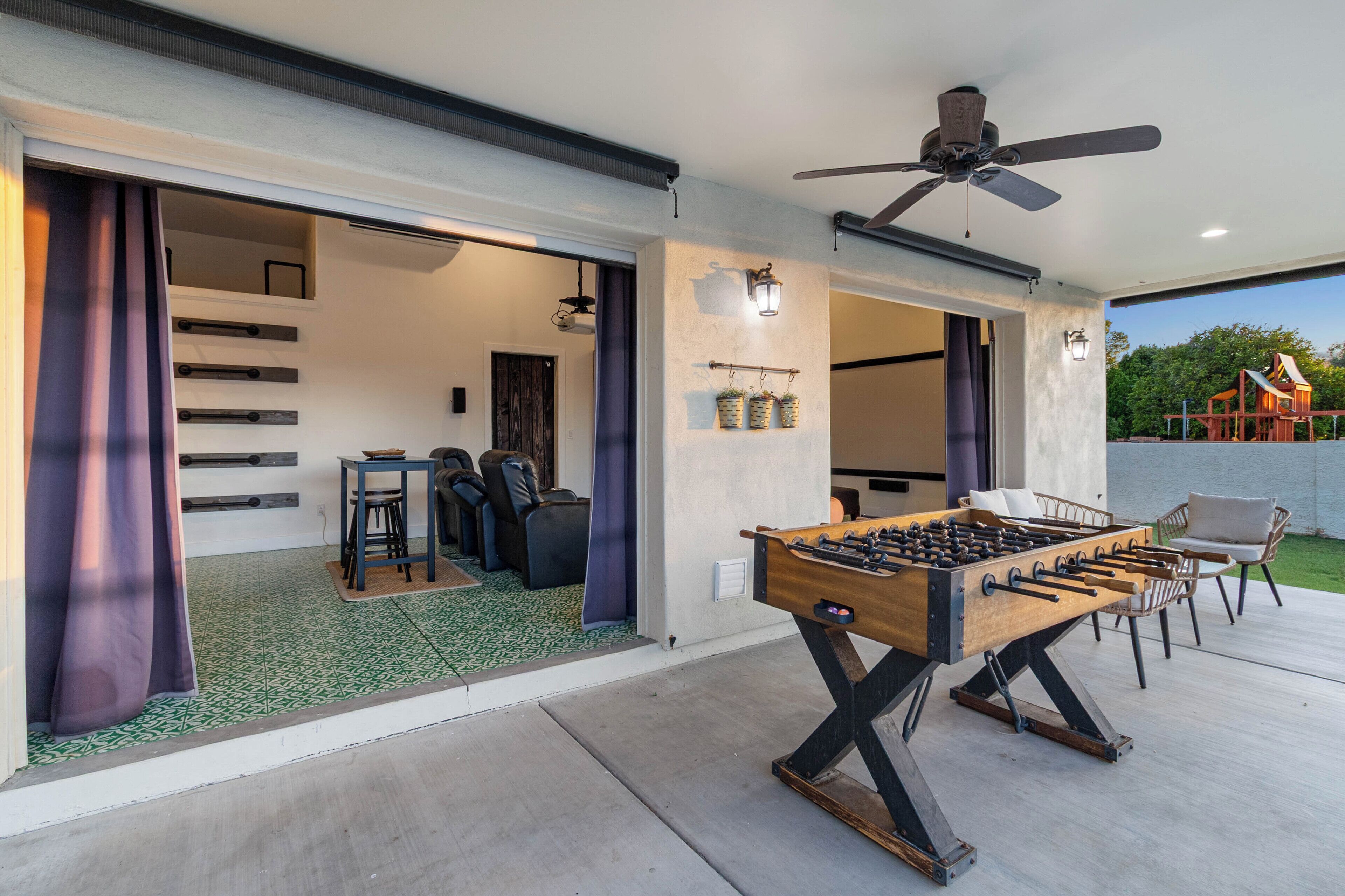 A modern indoor/outdoor living space with a foosball table, seating area, and a ceiling fan.