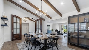 Uma sala de jantar com uma grande mesa de madeira, cadeiras pretas e um candeeiro. Há um armário de madeira com portas de vidro e uma porta branca com painéis de vidro.