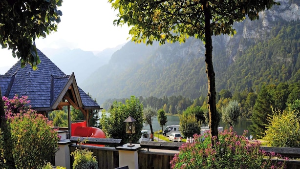 Ein Sitzbereich im Freien mit Tischen und Stühlen, einem Grill und Blick auf einen See und Berge im Hintergrund.