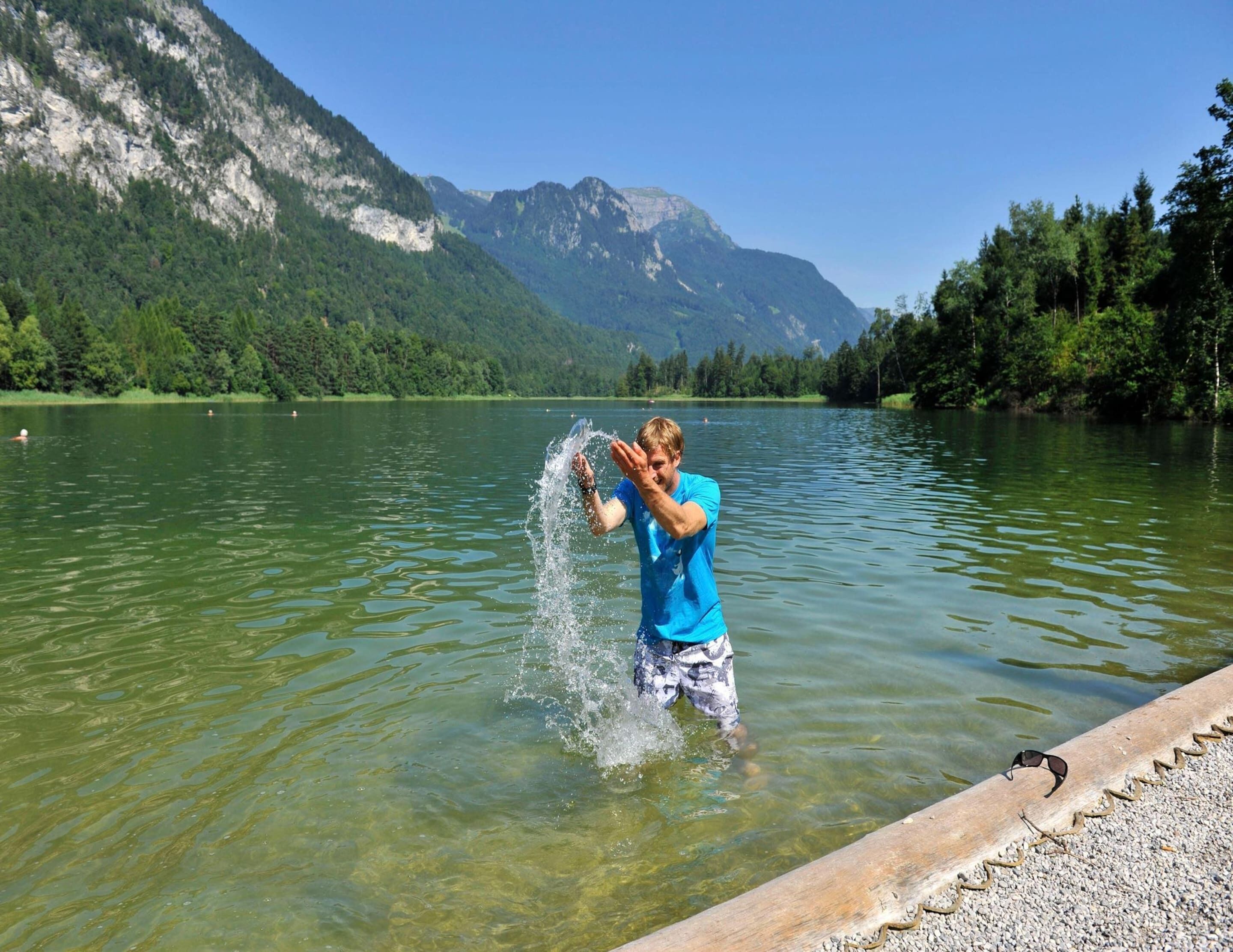 Eine Person mit blauem Hemd planscht in einem See vor einer Bergkulisse.