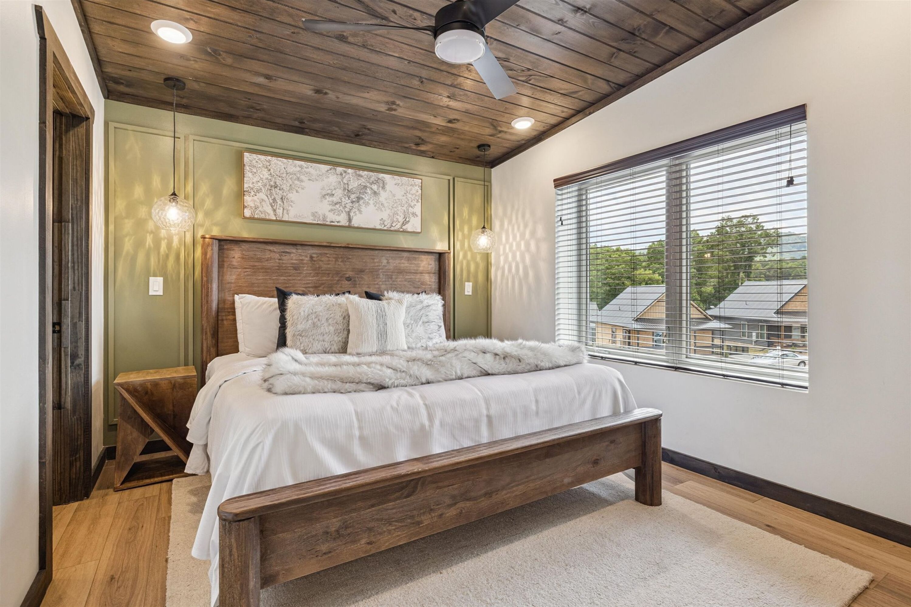 A bedroom with a wooden ceiling, a large bed with white bedding, a wooden bench, and a window with blinds.