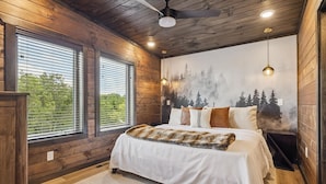 Ein Schlafzimmer mit Holzbalkendecke, großen Fenstern, einem Bett mit Felldecke und einem Wandgemälde von einem Wald.