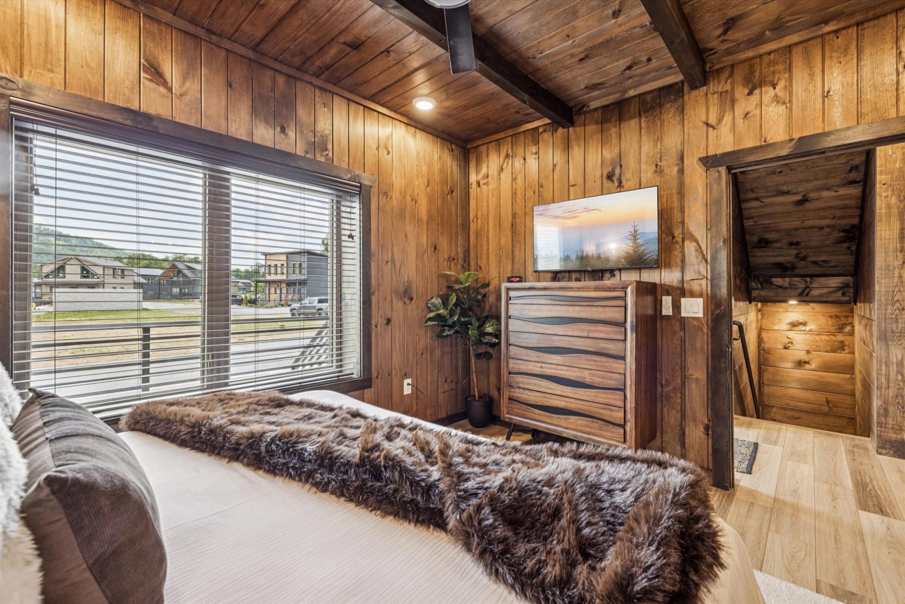 Ein Zimmer mit Holzwänden, einem großen Fenster mit Jalousien, einem Bett mit flauschigem Teppich, einer Pflanze und einem Bild an der Wand.