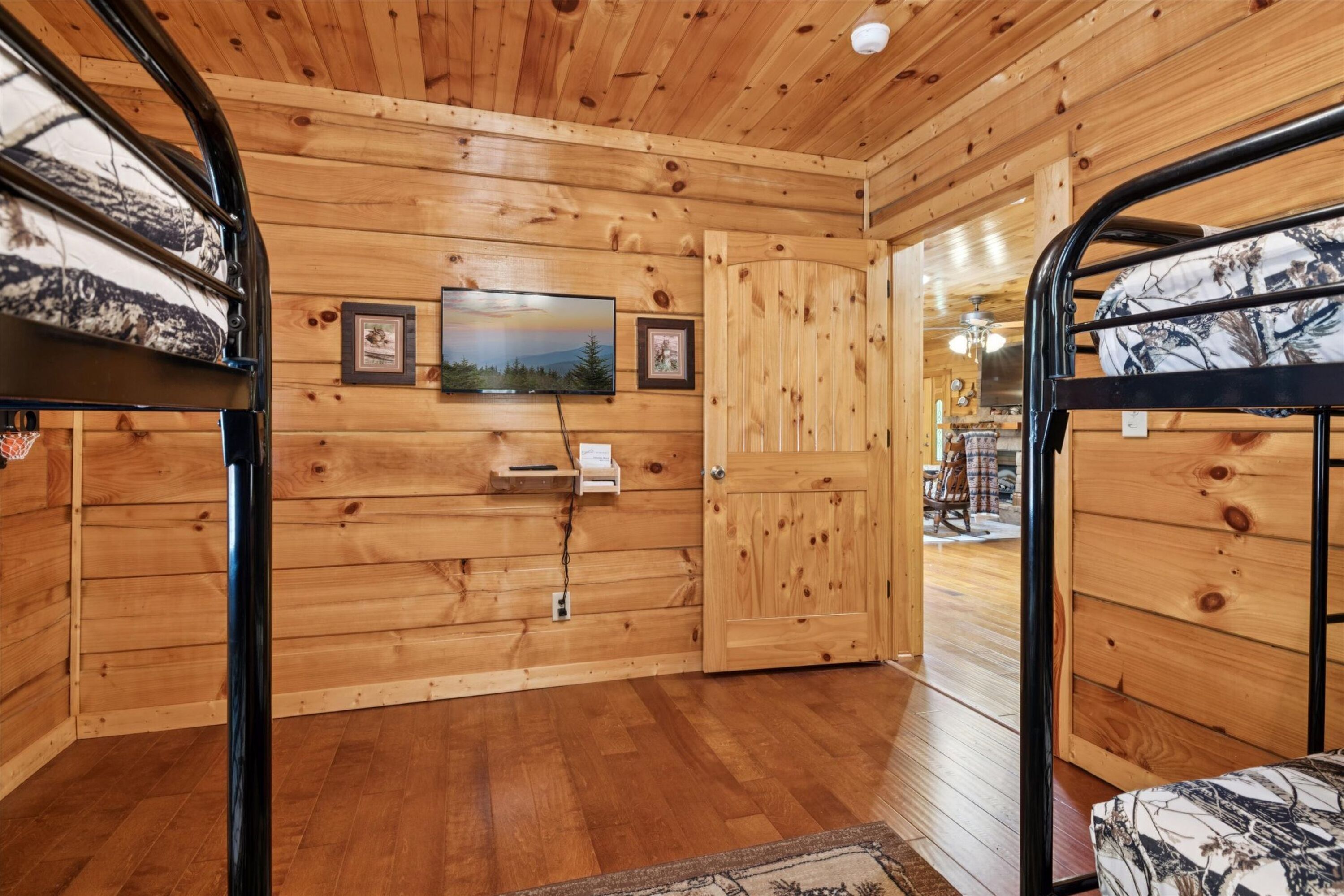 A wooden cabin interior with a television mounted on the wall, a door leading outside, and a bed with a patterned cover.
