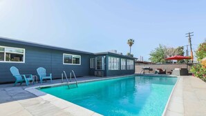 A rectangular swimming pool with a tiled deck, two Adirondack chairs, and a red umbrella. - Your Home Away From Home in the Sought Historic Huntridge Hills Neighborhood (Las Vegas)