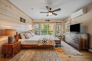 A bedroom with a wooden floor, a bed with a quilt, a ceiling fan, a dresser, a TV, and a window with a view of trees. - Tuckamore | Private Waterfront on Beaver Mountain Lake | Hot Tub (Rangeley)