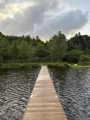 Un muelle de madera que se adentra en un lago, rodeado de un bosque frondoso y un edificio al fondo.