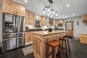A modern kitchen with wooden cabinets, stainless steel appliances, a central island with stools, and a large window. - Tuckamore | Private Waterfront on Beaver Mountain Lake | Hot Tub (Rangeley)