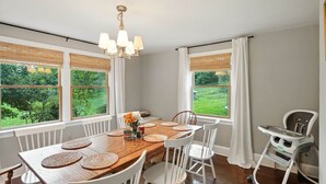 A dining area with a wooden table, white chairs, a chandelier, and large windows with views of greenery.