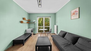 A modern living room with a sofa, a coffee table, a small table, and a floor lamp. There is a window with a view of trees and a radiator on the wall. - Grzegórzki Comfort Apartment (Kraków)