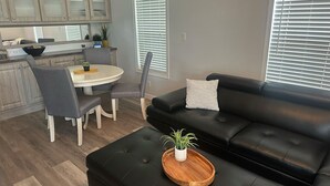 A modern living room with a black leather sofa, a dining table with chairs, and a kitchen area with cabinets and a window.