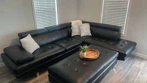 A black leather sectional sofa with a wooden tray and a plant, against a grey wall with blinds.