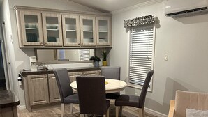 A compact kitchen with wooden cabinets, a dining table with four chairs, and a small seating area with a sofa and a cushion.