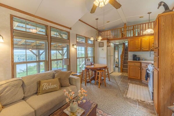 A spacious living room with a sofa, a coffee table, and a dining area with a table and chairs. There is a kitchen area with wooden cabinets and a staircase leading to an upper level.