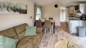 A cozy living room with a brown sofa, a dining table with chairs, a kitchen area with a microwave, and a clock on the wall.