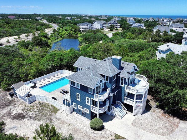 A large, multi-story house with a swimming pool, surrounded by lush greenery and a lake.