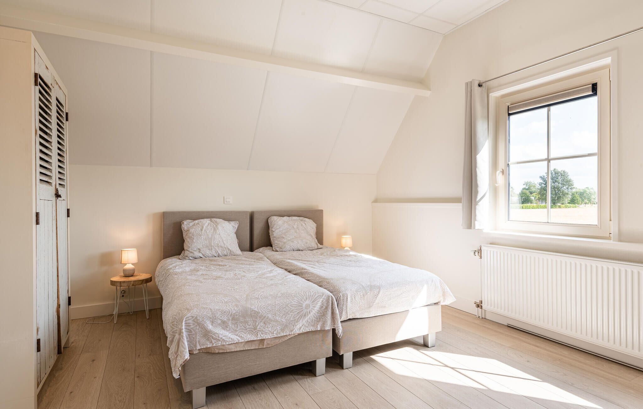 Een slaapkamer met een schuine kap, een groot raam, een radiator en een nachtkastje met een lamp.