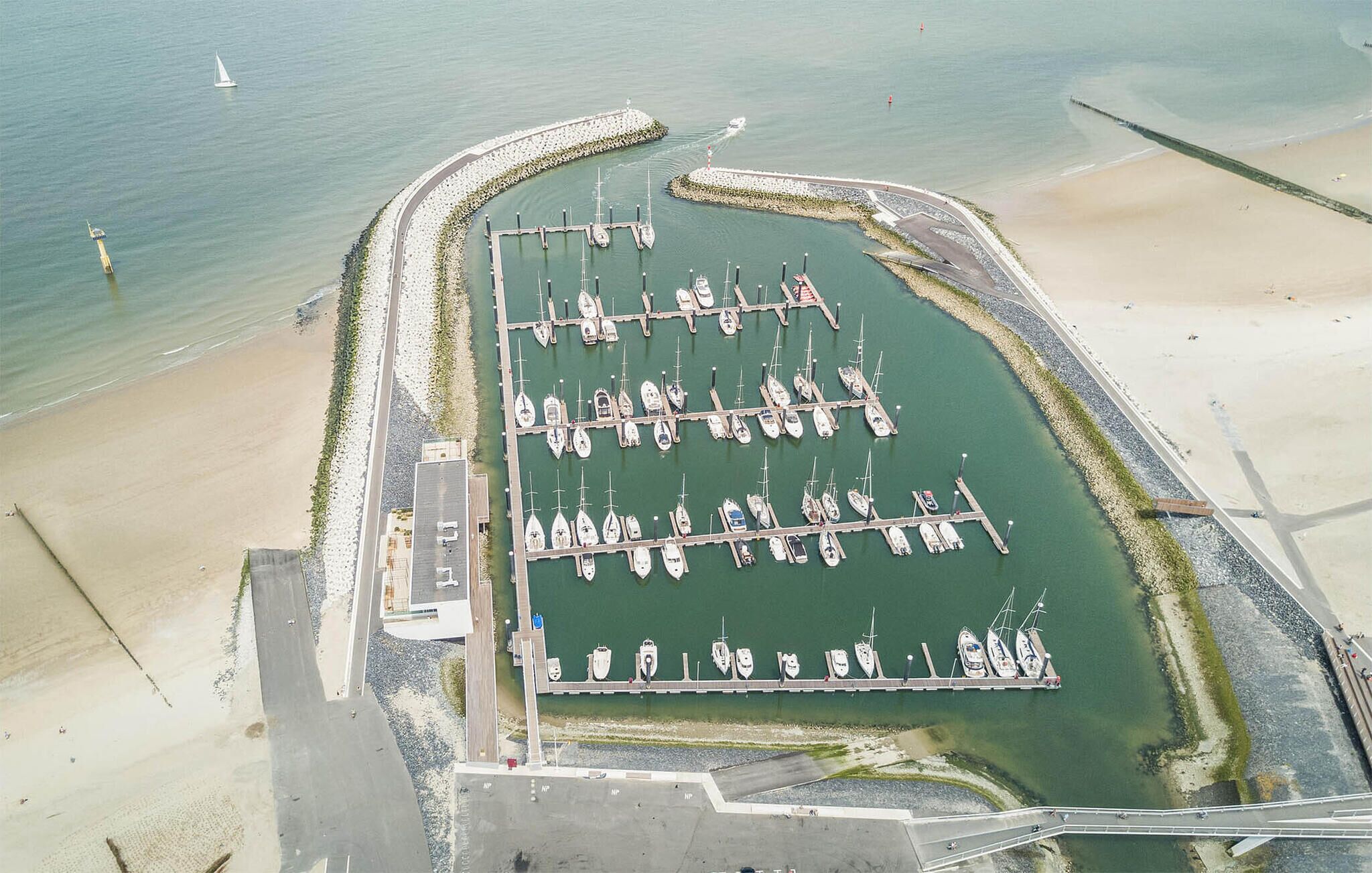 Vista aérea de un puerto deportivo con varios muelles y barcos, rodeado de una playa de arena y una barrera protectora.