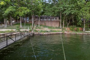A riverside property with a wooden deck, a covered outdoor area, and a building with a modern design. - Quiet Cove, Private Dock, Fishing, Swimming, Boat Slip & Kayaks (Camdenton)