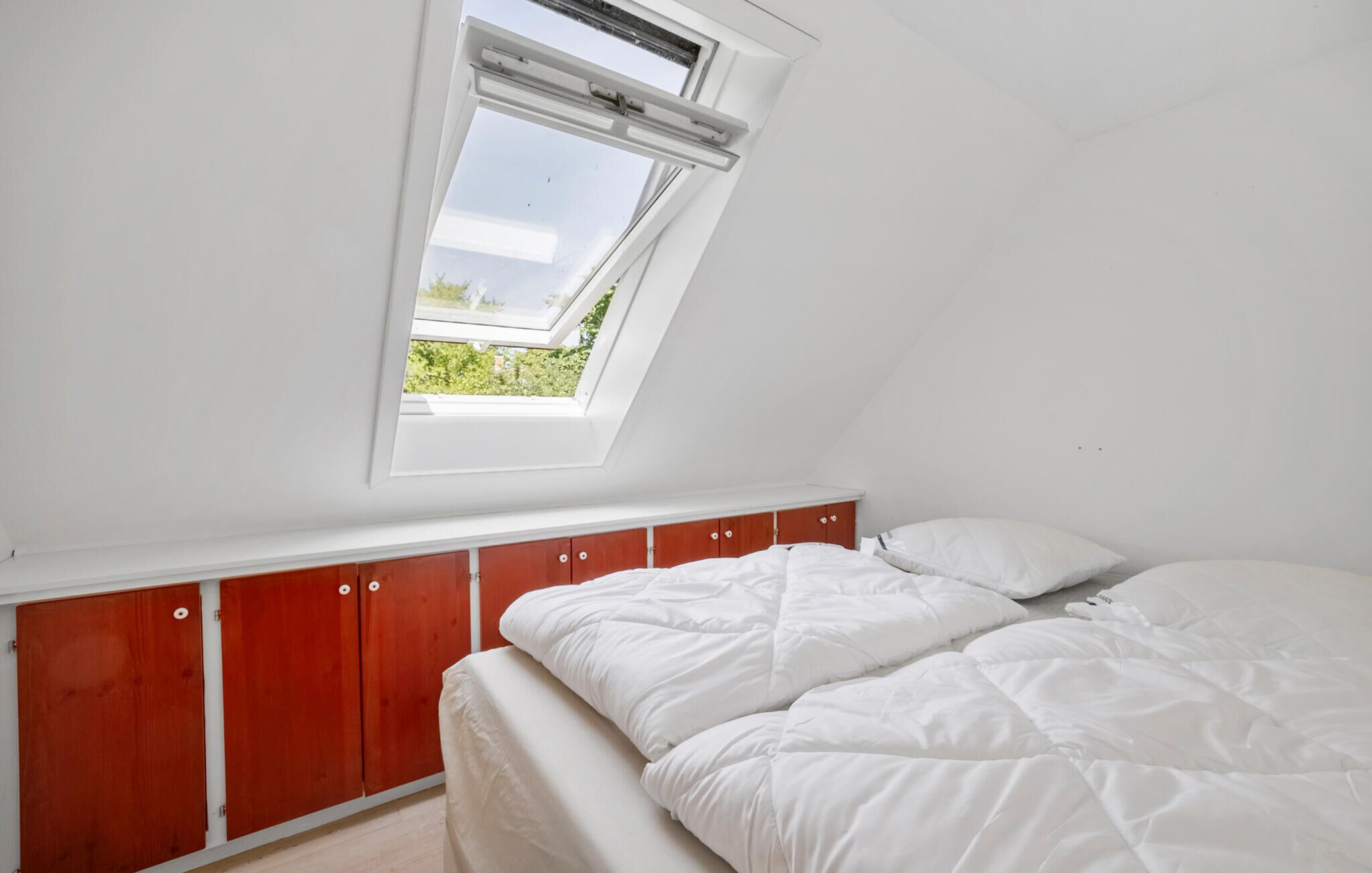 A bedroom with a sloped ceiling, a skylight, a bed with white bedding, and wooden cabinets below the window.