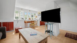 A living room with a wooden coffee table, a TV on a stand, a dining area with a table and chairs, and a kitchen with cabinets and a window. - Nice home in Svaneke with WiFi (Svaneke)