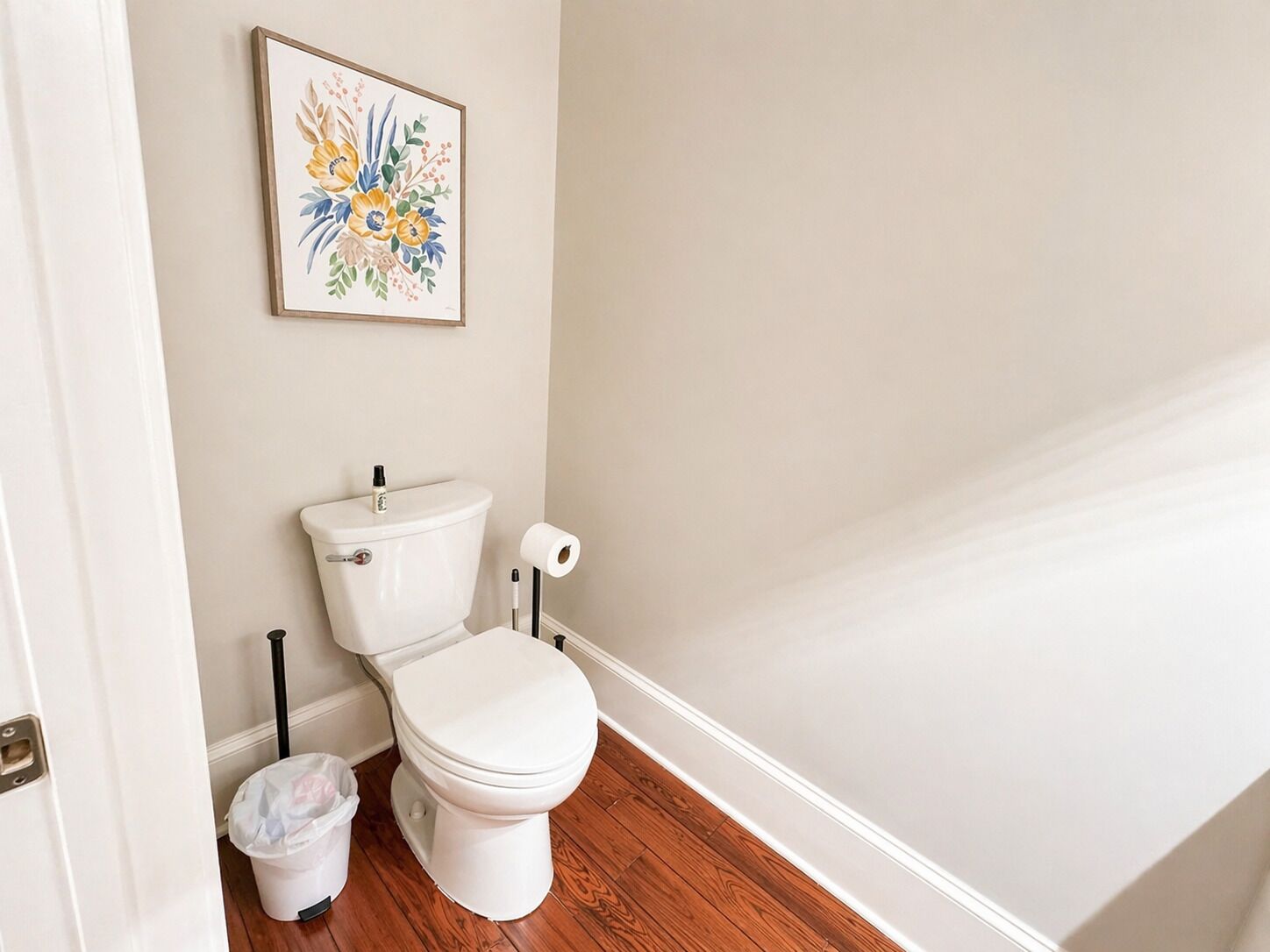 Een badkamer met een toilet, een rol wc-papier, een prullenbak en een lijst met bloemenkunst aan de muur.